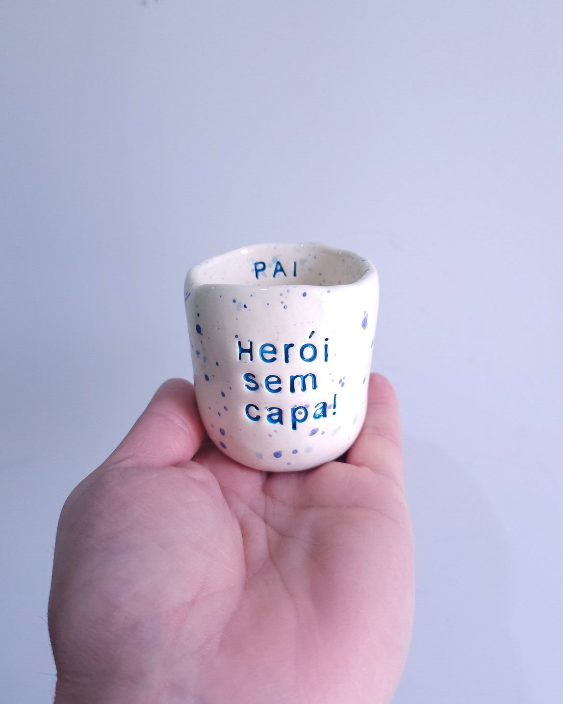 Copo de café em cerâmica artesanal branca com salpicado azul e a frase "Pai Herói sem capa" gravada à mão