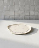 Bandeja de cerâmica artesanal branca com formato orgânico, corações lilás e a frase 'Lugar reservado para as merdas da mãe' escrita à mão na borda.