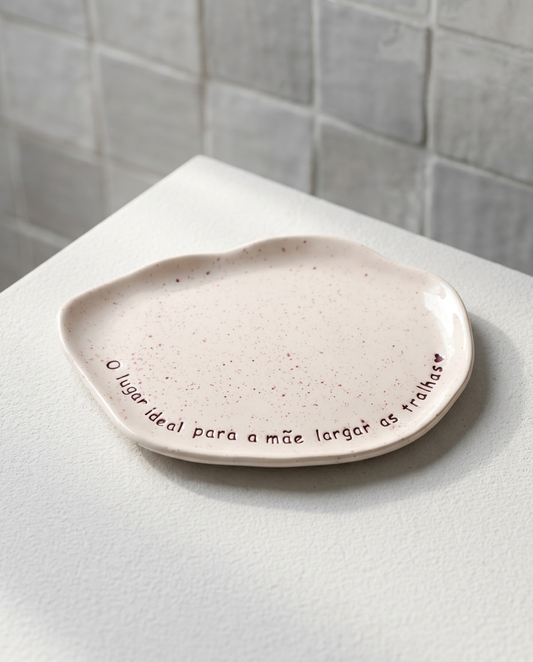Bandeja de cerâmica artesanal branca salpicada com a frase 'O lugar ideal para a mãe largar as tralhas' escrita à mão.