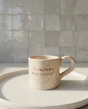 caneca de ceramica artesanal com pega salpicada com cores coloridas colocada sobre um prato branco com a frase eu reclamo mas resolvo marca tia cila