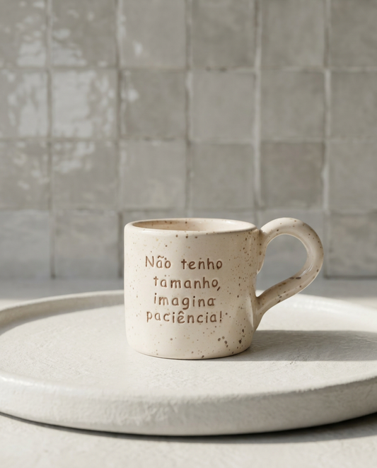 caneca de ceramica artesanal com pega salpicada em bege colocada sobre um prato branco com a frase nao tenho tamanho imagina paciencia marca tia cila