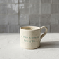 Caneca de cerâmica artesanal modelo Francisca, cor creme com salpicados verdes e turquesa. Ao centro, a frase "A tua caneca favorita..." gravada com um pequeno coração, fotografada com luz natural sobre superfície texturizada.