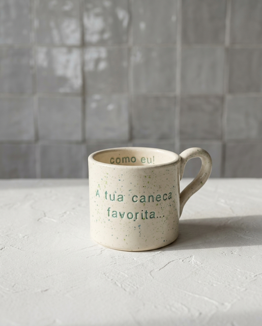 Caneca de cerâmica artesanal modelo Francisca, cor creme com salpicados verdes e turquesa. Ao centro, a frase "A tua caneca favorita..." gravada com um pequeno coração, fotografada com luz natural sobre superfície texturizada.