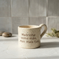 Caneca de cerâmica artesanal modelo Francisca, cor creme com salpicados ocre. Ao centro, a frase "Madrinha: como mãe, mas mais fixe!" gravada com um pequeno coração, fotografada com luz natural e pedras decorativas.