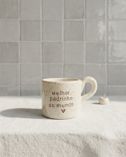 Caneca de cerâmica artesanal modelo Francisca, cor creme com salpicados coloridos e asa larga arredondada. Ao centro, a frase "melhor padrinho do mundo" gravada com um pequeno coração, fotografada com luz natural.
