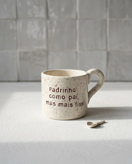 Caneca de cerâmica artesanal modelo Francisca, cor creme com salpicados ocre. Ao centro, a frase "Padrinho como pai, mas mais fixe!" gravada com um pequeno coração, fotografada com luz natural e uma colher pequena.