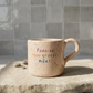 Caneca de cerâmica artesanal com asa (Modelo Francisca) de cor bege com salpicos coloridos, personalizada com a frase "Foda-se, sou granda mãe!" em letras coloridas, sobre uma base de pedra.