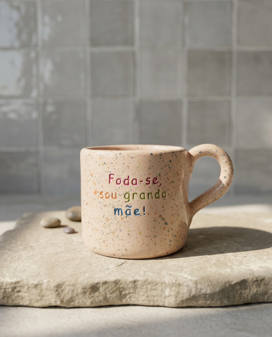 Caneca de cerâmica artesanal com asa (Modelo Francisca) de cor bege com salpicos coloridos, personalizada com a frase "Foda-se, sou granda mãe!" em letras coloridas, sobre uma base de pedra.