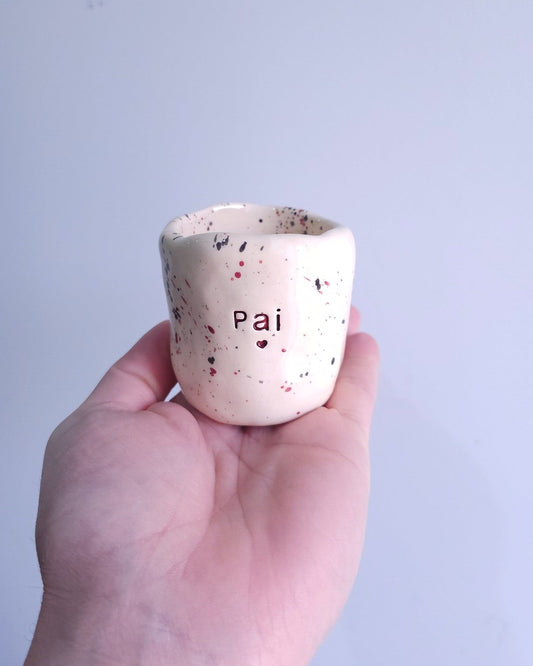 Copo de cerâmica artesanal Tia Cila com salpicado em bordeaux e preto, com a palavra "Pai" gravada na frente, seguro por uma mão.
