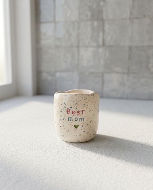 Copo de café artesanal sem asa (Modelo Inês) em cerâmica com salpicos coloridos, apresentando a frase "Best mom" em tons de rosa e azul com um coração verde gravado.