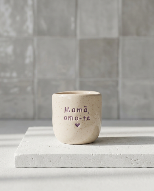 Copo de café artesanal sem asa, modelo Inês, em cerâmica salpicada com a frase "Mamã, amo-te" e um coração gravados em tom violeta, sobre uma base de pedra branca.