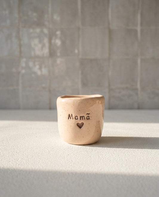 Copo de café artesanal sem asa (Modelo Inês) em cerâmica bege salpicada, com a palavra "Mamã" gravada e um pequeno coração vazado por baixo, iluminado pela luz solar.