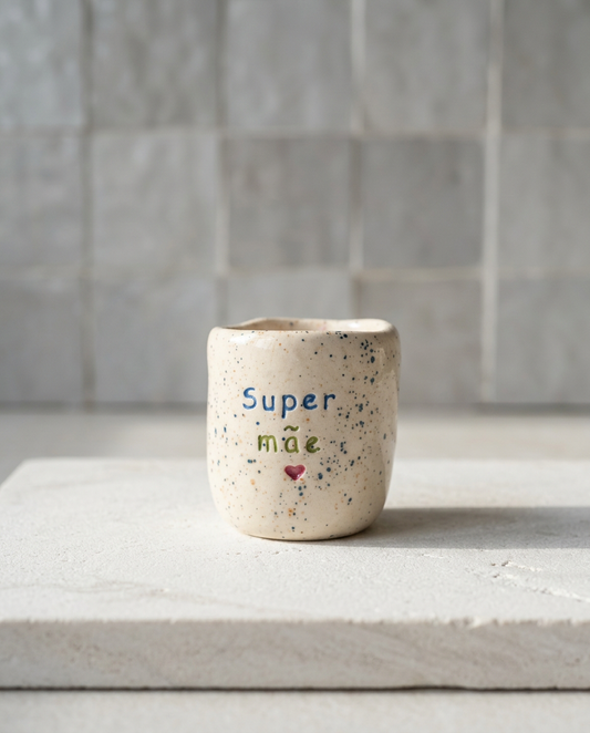 Copo de café artesanal sem asa (Modelo Inês) em cerâmica com salpicos azuis, apresentando a frase "Super mãe" gravada em azul e verde com um coração rosa.