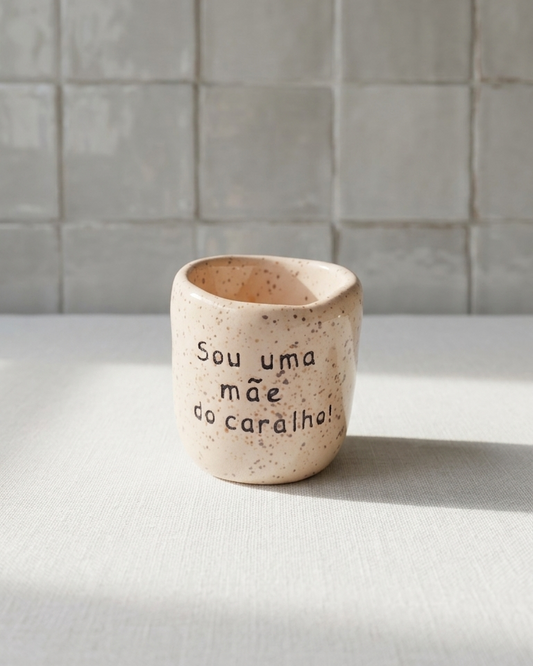 Copo de café artesanal sem asa (Modelo Inês) em cerâmica rosada salpicada com a frase gravada "Sou uma mãe do caralho!" e fumo a sair do interior, sobre tecido branco.