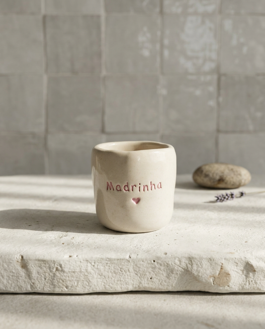 Copo de cerâmica artesanal modelo Inês, em tom creme com salpicados rosa pastel. No centro, a palavra "Madrinha" e um coração gravados em baixo relevo com cor rosa, sob luz solar natural.