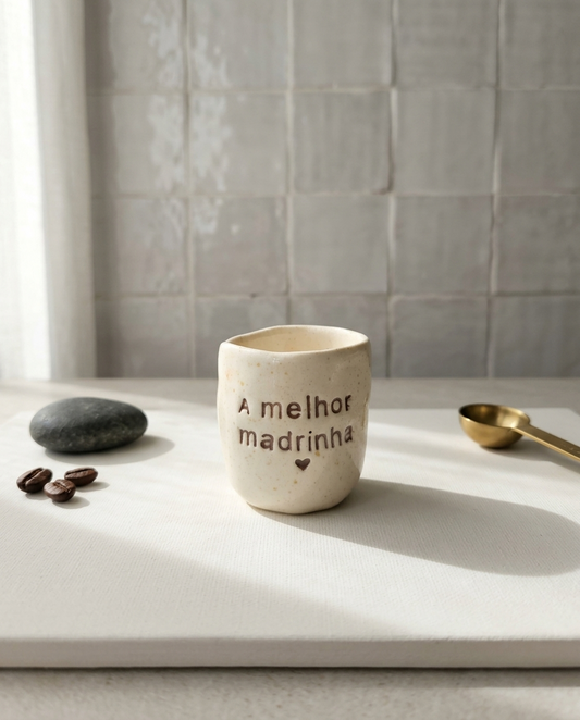 Copo de cerâmica artesanal modelo Inês, cor creme com salpicados ocre. Ao centro, a frase "A melhor madrinha" gravada em baixo relevo com um pequeno coração, fotografado com grãos de café e colher.