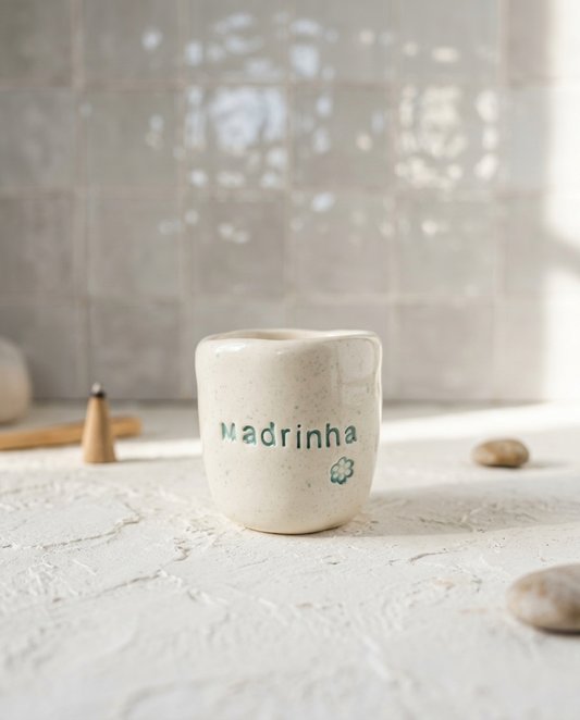 Copo de cerâmica artesanal modelo Inês, cor creme com salpicados verde água. No centro, a palavra "Madrinha" e uma pequena flor gravadas em baixo relevo na cor verde, sobre superfície texturizada.