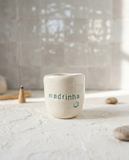 Copo de cerâmica artesanal modelo Inês, cor creme com salpicados verde água. No centro, a palavra "Madrinha" e uma pequena flor gravadas em baixo relevo na cor verde, sobre superfície texturizada.
