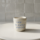 Copo de café artesanal modelo Inês, em cerâmica clara com salpicados azul suave. Ao centro, a frase gravada "café do melhor padrinho", fotografado sobre um prato de textura mate.