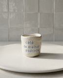 Copo de café artesanal modelo Inês, em cerâmica clara com salpicados azul suave. Ao centro, a frase gravada "café do melhor padrinho", fotografado sobre um prato de textura mate.