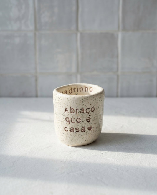 Copo de cerâmica artesanal modelo Inês, cor creme com salpicados verdes. Apresenta a frase "Abraço que é casa" gravada no exterior e a palavra "Padrinho" no rebordo interior.