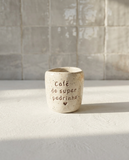 Copo de café artesanal modelo Inês, em cerâmica clara com salpicados verdes e laranjas. Ao centro, a frase gravada "café do super padrinho" com um pequeno coração, fotografado sob luz natural.