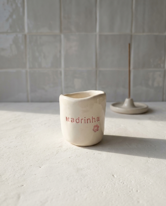 Copo de cerâmica artesanal modelo Inês, cor creme, decorado com flores em relevo pintadas em tons de rosa ao redor da palavra "Madrinha".