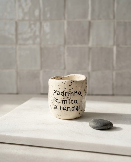 Copo de cerâmica artesanal modelo Inês, cor creme com salpicados escuros. Apresenta a frase "Padrinho, o mito, a lenda!" gravada no exterior, fotografado sobre uma placa de mármore.